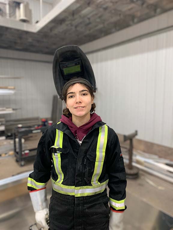 A female welder poses with her welder's helmet lifted to reveal her face.